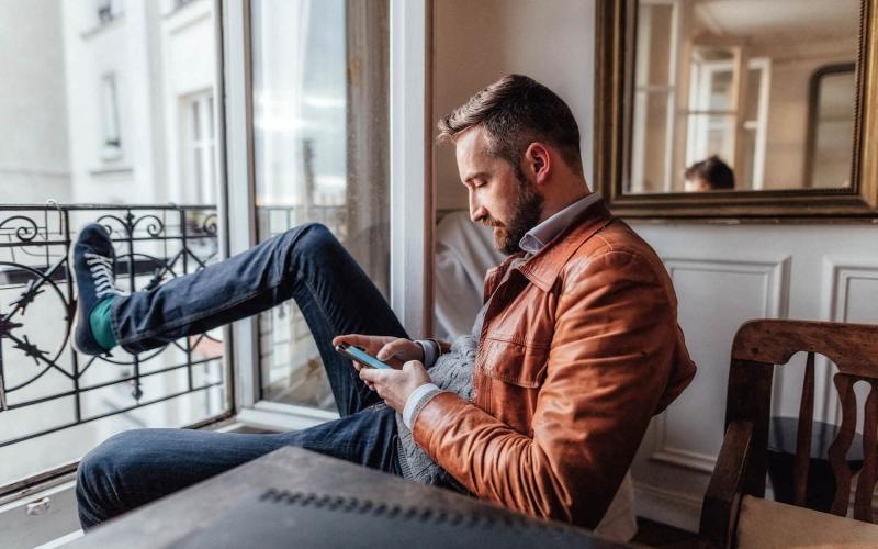 a man sits on a chair and rests his foot on a balcony railing as he looks at his phone in his room