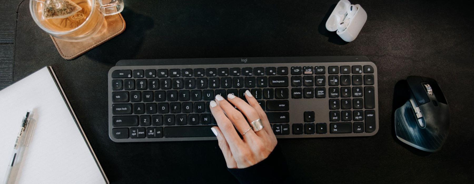 woman's hand on a keyboard surrounded by office items and a cup of tea on a coaster